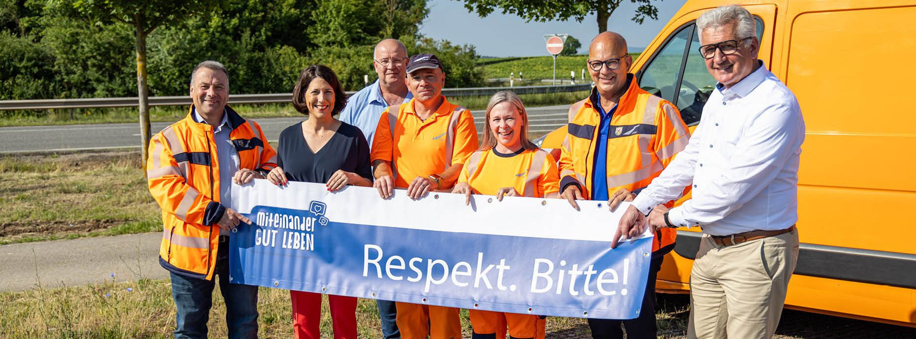 Die Ministerin Daniela Schmitt, der LBM-Geschäftsführer, zwei Streckenwarten und drei weitere Personen halten einen Banner fest. Auf dem Banner steht "Respekt. Bitte". Die Gruppe steht vor dem Auto der Streckenkontrolle.