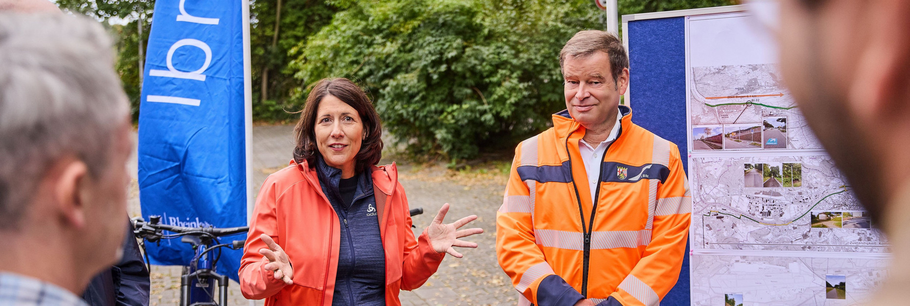 Verkehrsministerin Daniela Schmitt und stellvertretender LBM-Geschäftsführer bei der Pressekonferenz zu den Pendlerradrouten.  Hinter ihnen ist ein links ein Fahrrad und rechts eine Karte zu sehen. 