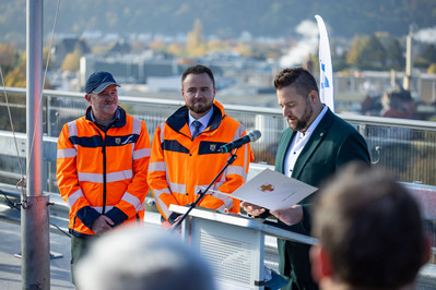 Ein Mann steht an einem Rednerpult und ließt eine Urkunde vor. Zwei Männer in orangenen Jacken warten daneben.