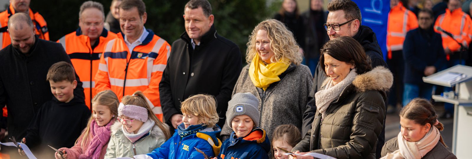 Verkehrsministerin Daniela Schmitt und mehrere Kinder schneiden mit Scheren ein weißes Band durch. Dahinter stehen der stellvertretende LBM-Geschäftsführer Lutz Nink mit oranger Jacke und weitere Vertreterinnen und Vertreter. 